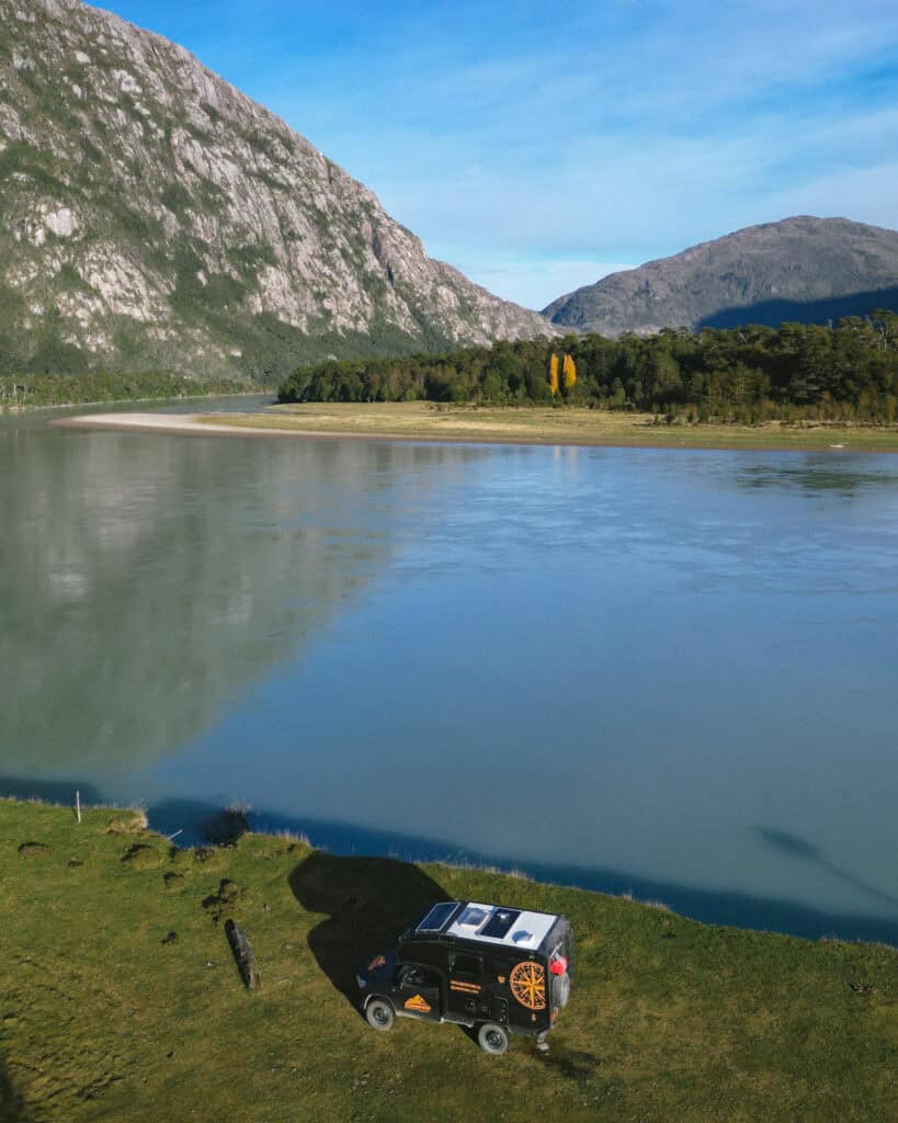 Aerial view of a campervan parked beside the calm waters of Río Baker in Caleta Tortel, Chile, with green forests, sandy riverbanks, and distant golden trees