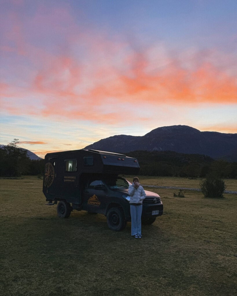 Our Epic 3-Week Carretera Austral Road Trip Itinerary 14 Sally stands bundled in a jacket next to a campervan under a vivid pink and orange sunrise sky in Caleta Tortel, Chile, with mountains silhouetted in the distance