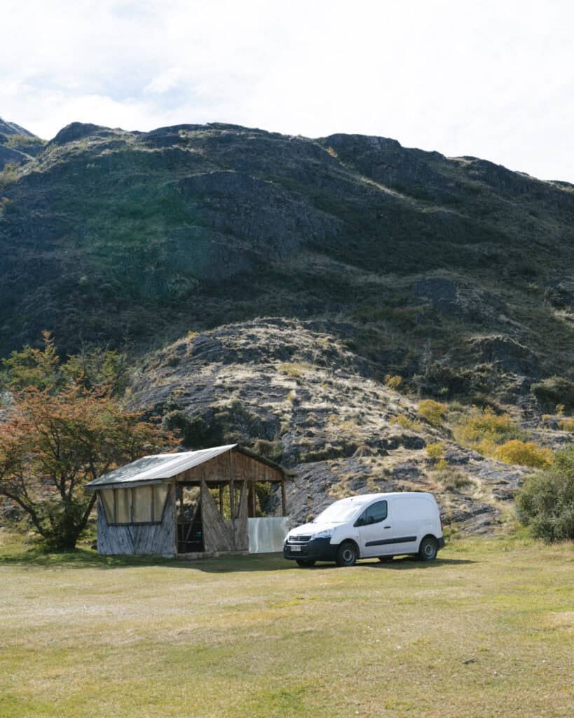 Camping on the Carretera Austral, Chile: Our Tips & Tricks 13 A rustic wooden shelter and white van sit on a grassy patch at the base of rocky hills in Parque Nacional Patagonia, Tamango sector, near Cochrane, Chile