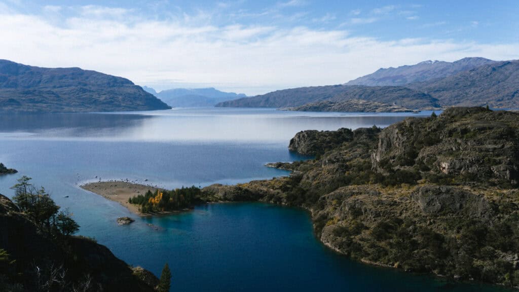 Valle Chacabuco Sector Guide: Parque Nacional Patagonia, Chile 42 Wide panoramic view of Lago Cochrane with glassy blue waters stretching between rugged mountains, a small forested peninsula visible in the foreground
