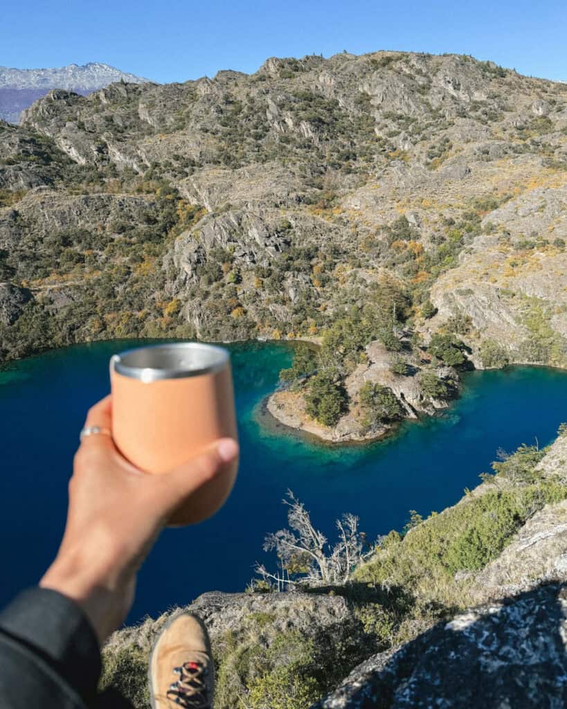 Tamango Sector Guide: Parque Nacional Patagonia, Chile 23 Sally’s hand holding a peach-colored cup while sitting on a cliff edge overlooking the turquoise and deep blue water of Lago Cochrane, surrounded by rocky ridges