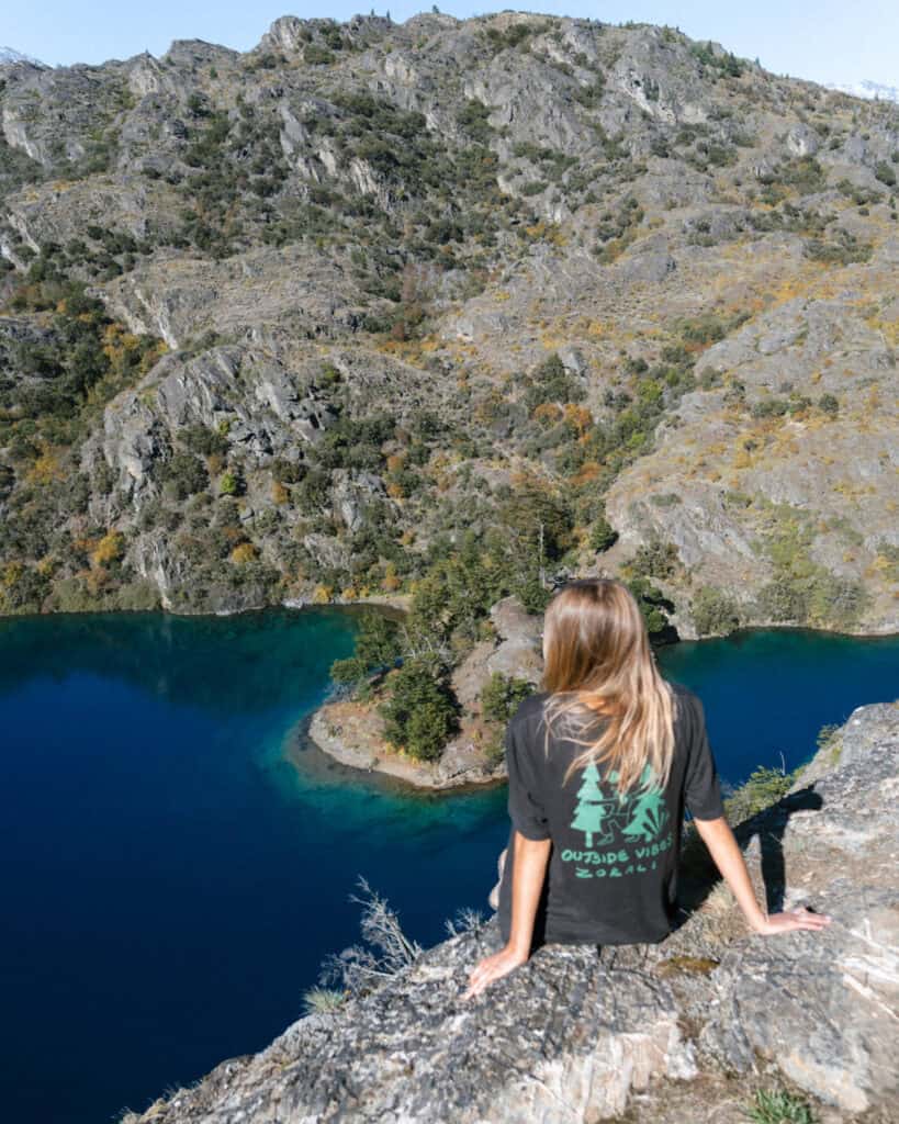 The Carretera Austral, Chile: Everything You Need to Know 48 Back view of Sally seated on a rocky outcrop overlooking a bright blue lake and forested island, surrounded by steep gray hills in Parque Nacional Patagonia near Cochrane