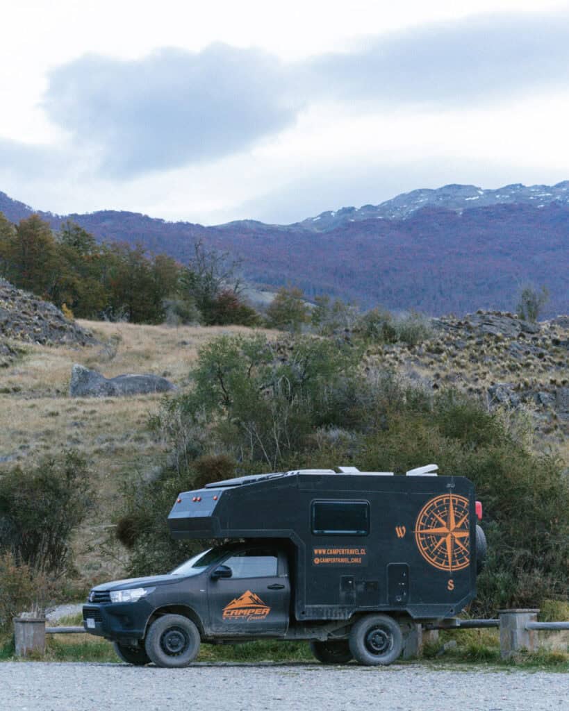 Villa Cerro Castillo, Chile Travel Guide: Hiking & Huemuls 8 A black camper truck parked on a gravel area beside autumn-colored trees, with mountain ridges in the distance under a cloudy sky