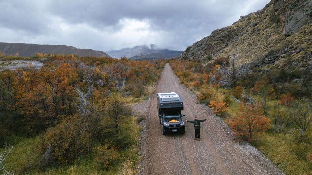 The Carretera Austral, Chile: Everything You Need to Know 71 Drone shot of Brayden standing with arms wide next to a black camper van on a gravel road surrounded by autumn foliage and mountainous terrain