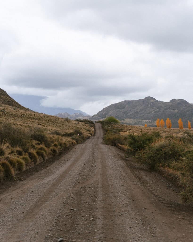 Valle Chacabuco Sector Guide: Parque Nacional Patagonia, Chile 11 A winding dirt road cuts through dry, rugged hills in Valle Chacabuco, Chile, with distant orange-leaved trees providing a vivid contrast against the overcast sky and mountainous horizon