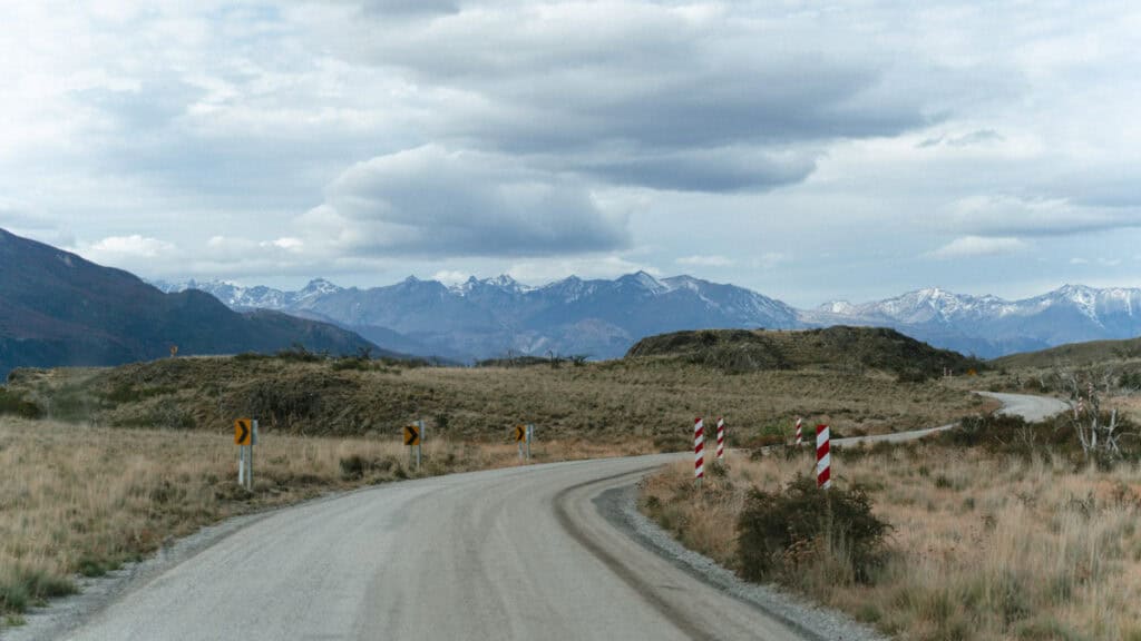 Valle Chacabuco Sector Guide: Parque Nacional Patagonia, Chile 4 Curving gravel road with red-and-white and yellow arrow roadside markers, winding through golden fields and leading toward snow-capped Andes mountains under a cloudy sky, captured in Sector Valle Chacabuco on Chile's Carretera Austral