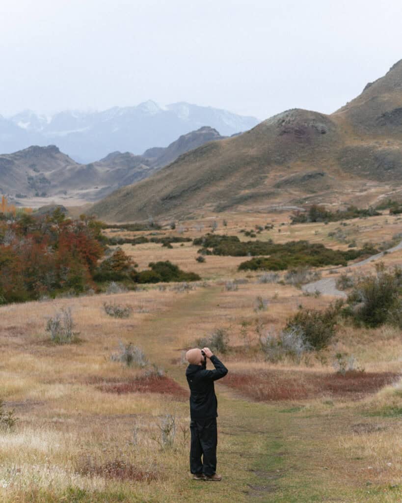 Valle Chacabuco Sector Guide: Parque Nacional Patagonia, Chile 38 Brayden, dressed in a black jacket and beige beanie, stands on a grassy trail in Sendero La Vega, looking through binoculars at the rugged hills and distant snowy peaks of Sector Valle Chacabuco, Parque Nacional Patagonia, Chile