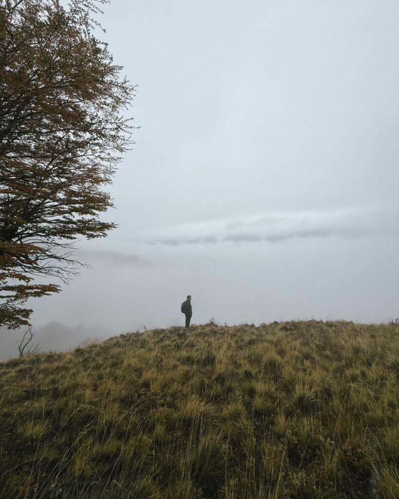 Valle Chacabuco Sector Guide: Parque Nacional Patagonia, Chile 31 Brayden stands on a grassy ridge surrounded by fog, facing the vast cloudy expanse along Sendero Lagunas Altas in Valle Chacabuco