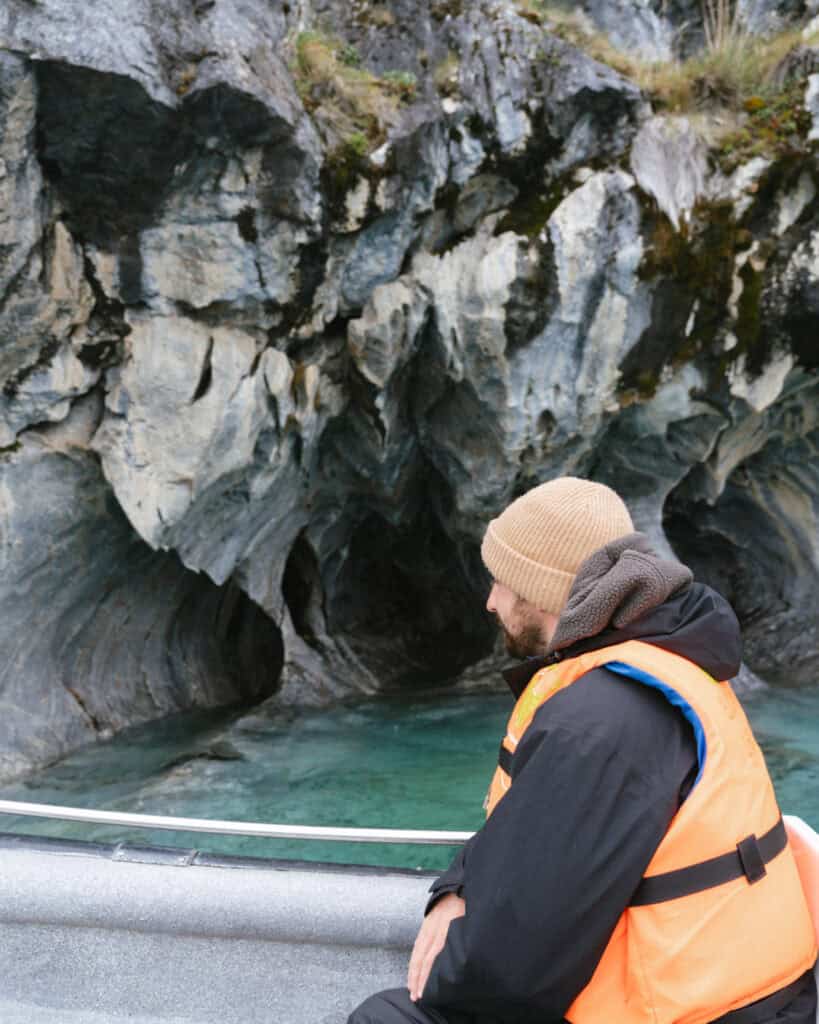 How to Visit the Marble Caves in Chile: Worth the Journey? 15 Brayden sits on a boat wearing an orange life vest and beige beanie, gazing at the rippled marble walls of the Capillas de Mármol rising from turquoise water in Puerto Río Tranquilo, Chile