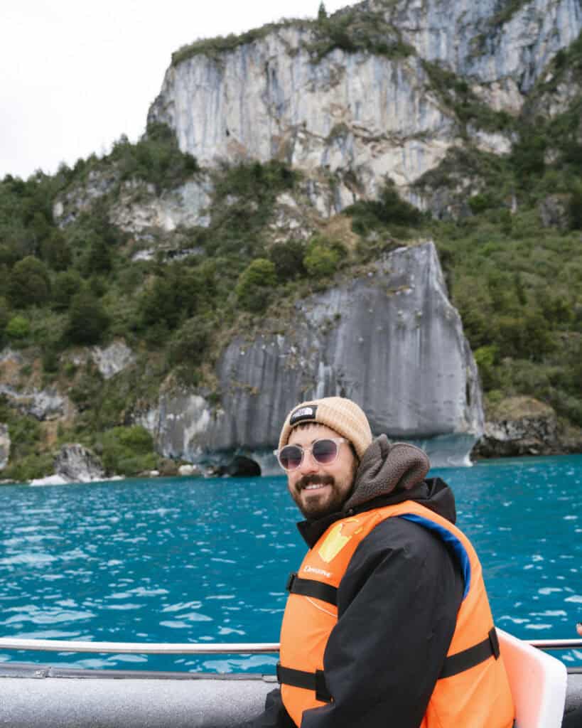 How to Visit the Marble Caves in Chile: Worth the Journey? 2 Brayden smiles at the camera from a boat on the vivid blue waters of Puerto Río Tranquilo, with the towering marble cliffs of the Capillas de Mármol behind him