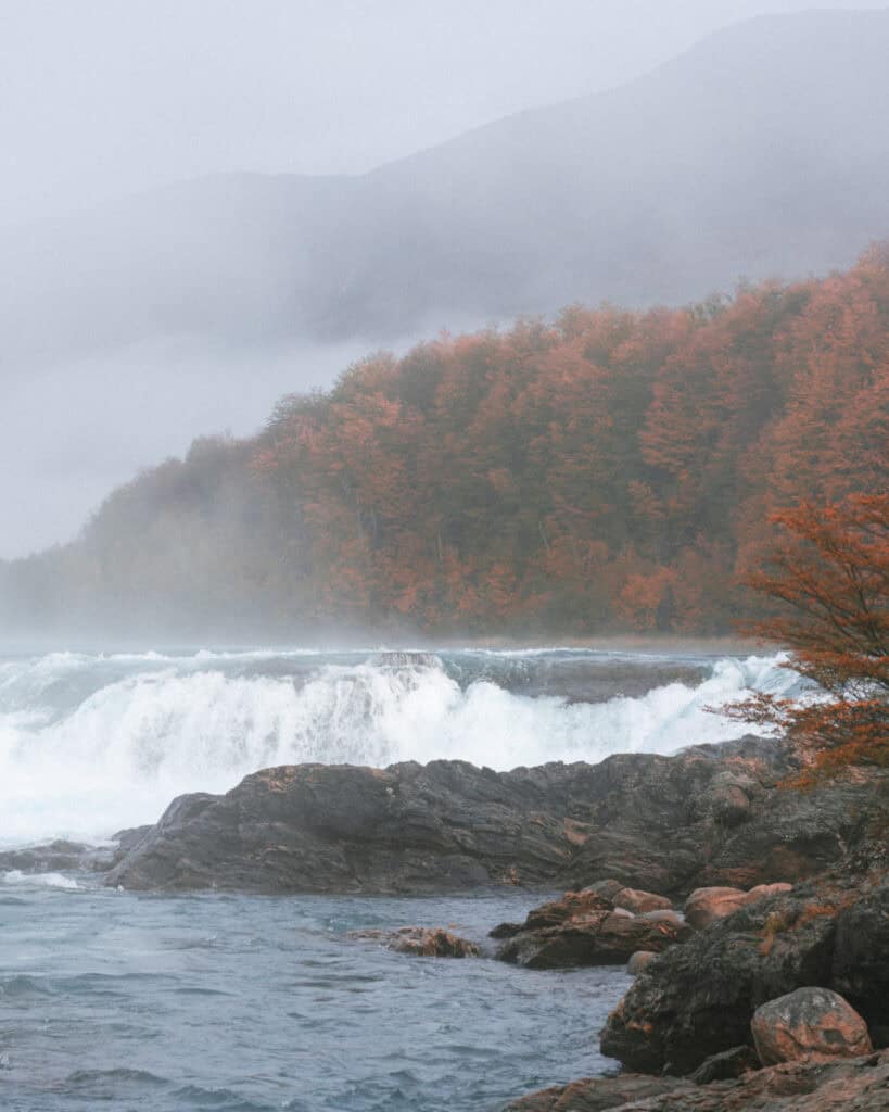 Puerto Rio Tranquilo, Chile Travel Guide: Marble & Ice 37 A foggy view of a cascading section of Río Baker with a forest of orange autumn foliage and hazy mountains in the background, near Puerto Río Tranquilo