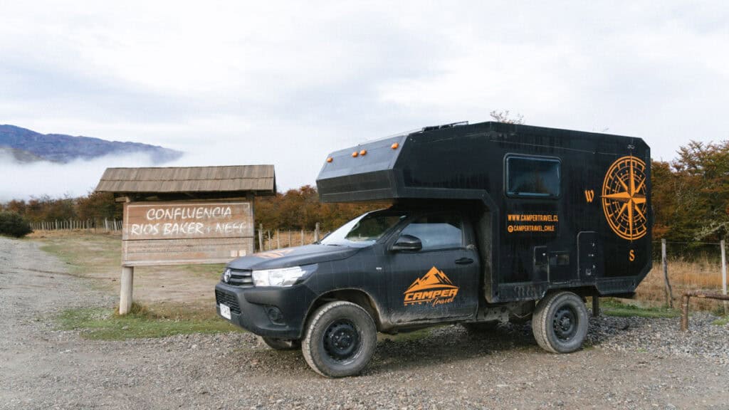 Puerto Rio Tranquilo, Chile Travel Guide: Marble & Ice 38 A black camper vehicle from Camper Travel Chile is parked beside a wooden sign reading “Confluencia Ríos Baker y Neff,” with autumn trees and misty mountains in the background