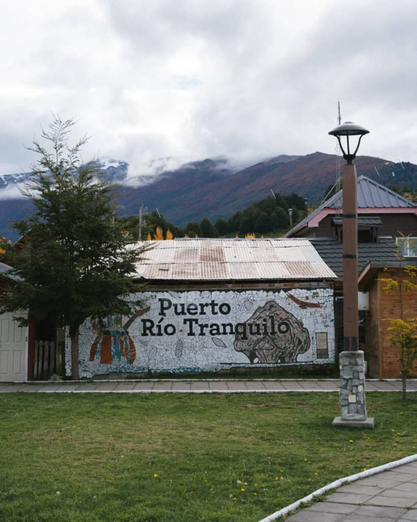 How to Visit the Marble Caves in Chile: Worth the Journey? 11 A colorful mosaic mural in the plaza of Puerto Río Tranquilo displays the town's name along with local imagery, set against a corrugated metal building and backed by cloudy mountains