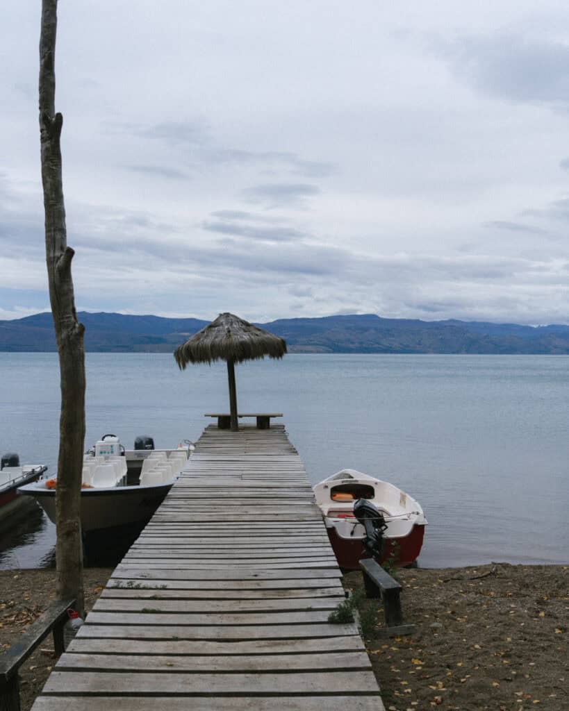 How to Visit the Marble Caves in Chile: Worth the Journey? 18 A narrow wooden pier extends over a serene lake, flanked by small boats and a single thatched umbrella at the end, with soft mountains and cloudy skies in the distance
