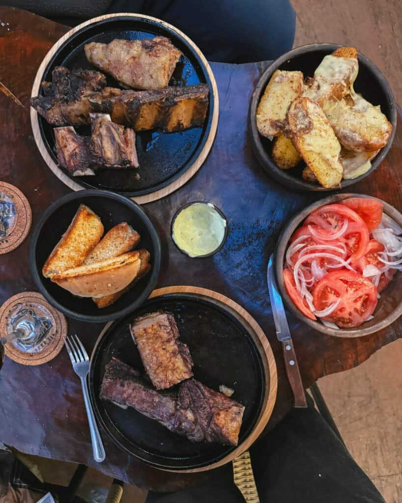 Puerto Rio Tranquilo, Chile Travel Guide: Marble & Ice 39 Overhead view of a hearty meal featuring grilled beef ribs, cheesy potato wedges, bread, and a tomato-onion salad, all served on rustic wooden platters and bowls
