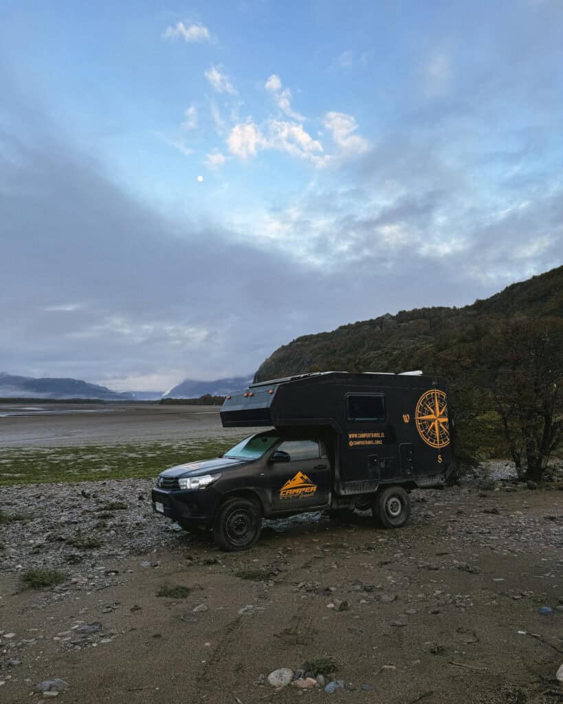 Hornopiren, Chile Travel Guide: Termas, Alerces & Fjords 10 A rugged black camper truck from "Camper Travel Chile" is parked on rocky terrain near a riverbed in Villa Cerro Castillo, with dramatic hills and a cloudy blue sky in the background
