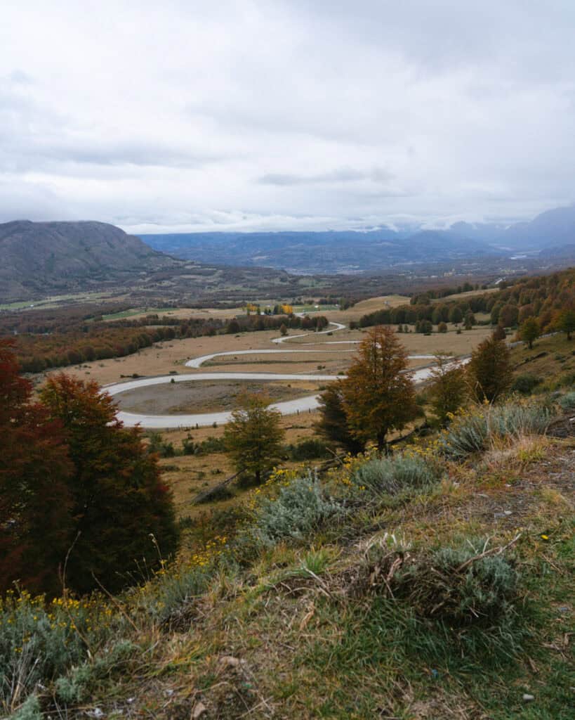 Villa Cerro Castillo, Chile Travel Guide: Hiking & Huemuls 22 A sweeping view from the Cuesta del Diablo in Villa Cerro Castillo National Park shows a winding road through a wide valley, with fall-colored trees and distant mountain ranges under a moody sky
