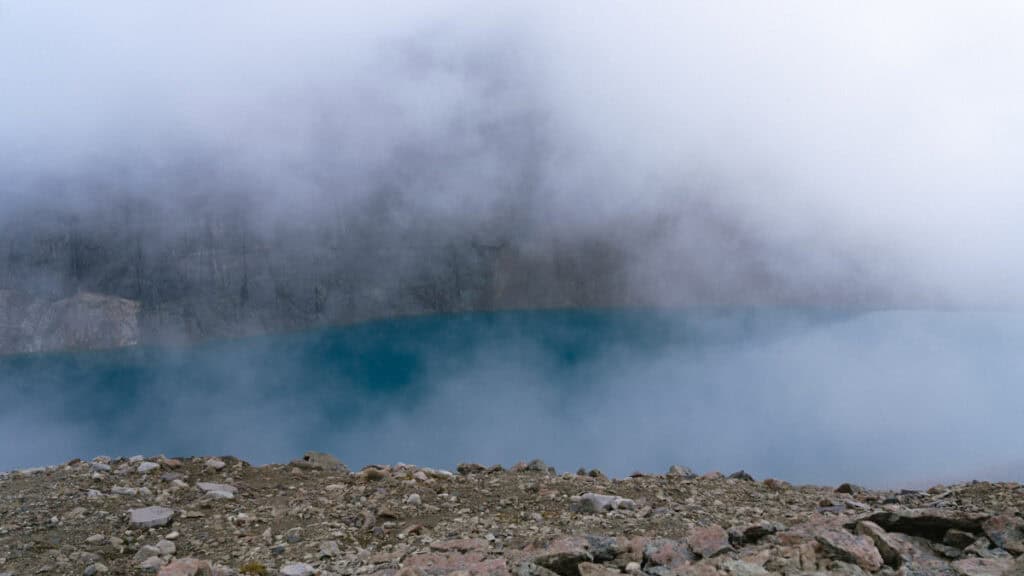 Laguna Cerro Castillo, Chile Hiking Guide 6 A panoramic view of a turquoise lagoon shrouded in fog at the base of a rocky mountain wall in Villa Cerro Castillo National Park, with scattered rocks in the foreground