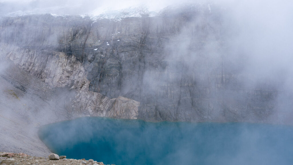 Laguna Cerro Castillo, Chile Hiking Guide 28 A cliff face runs along the edge of a bright blue lagoon in Villa Cerro Castillo National Park, with swirling clouds creating a mysterious and remote mood
