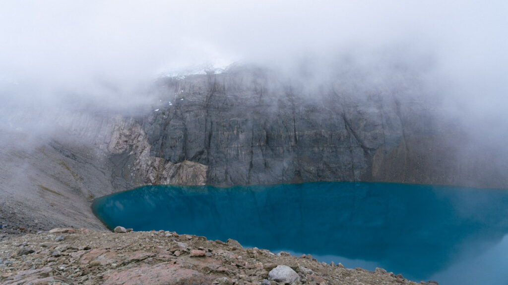 Villa Cerro Castillo, Chile Travel Guide: Hiking & Huemuls 27 A vivid turquoise lagoon sits beneath a towering, fog-draped cliff in Villa Cerro Castillo National Park, with jagged rocks and mist swirling at the water’s edge