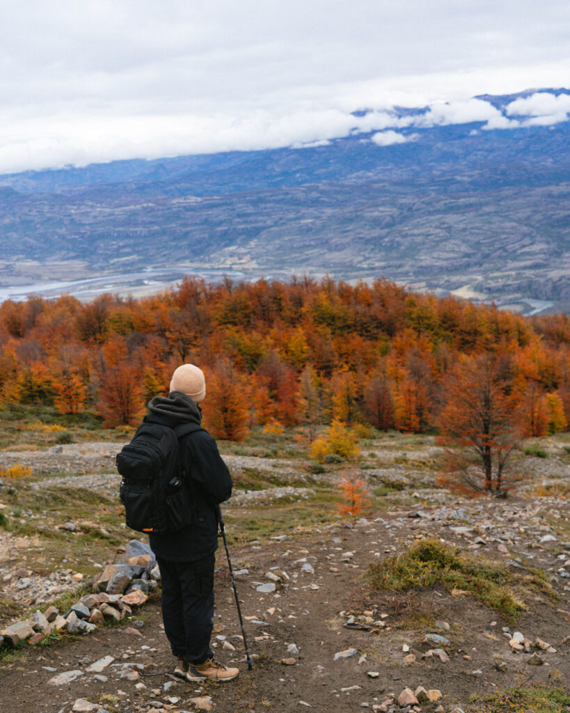 Villa Cerro Castillo, Chile Travel Guide: Hiking & Huemuls 17 Brayden stands with trekking poles on a rocky trail overlooking a forest of vibrant autumn colors in Villa Cerro Castillo National Park, with layered mountains fading into the distance