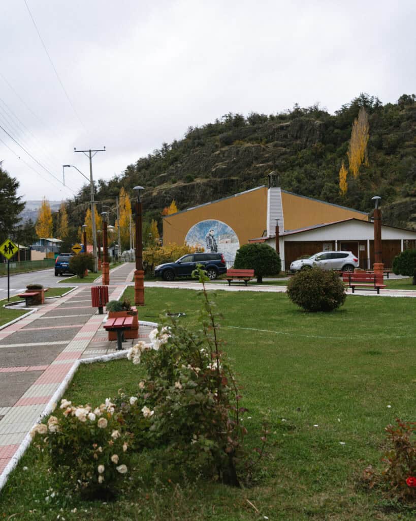Villa Cerro Castillo, Chile Travel Guide: Hiking & Huemuls 18 A park in the town of Villa Cerro Castillo, Chile, shows a sidewalk lined with benches and a mural on a tan building, framed by mountains and autumn trees