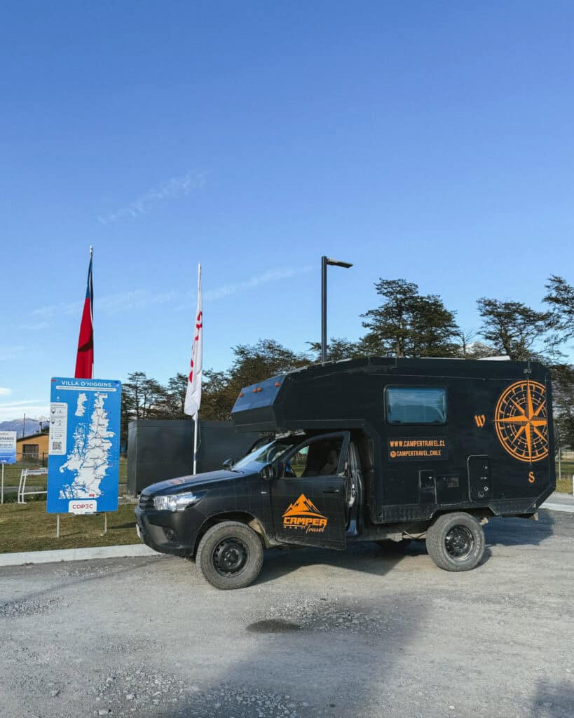 Villa O'Higgins, Chile Guide: End of the Carretera Austral 34 The Camper Travel Chile van is parked next to a blue Villa O'Higgins welcome sign and Chilean flags at a COPEC station, with a clear blue sky above and tall trees in the background