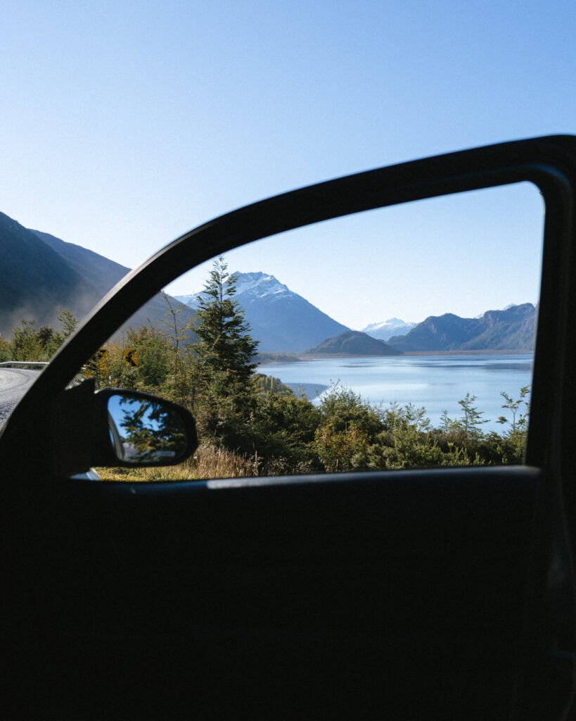 Villa O'Higgins, Chile Guide: End of the Carretera Austral 10 Looking through an open car door at a stunning Patagonian lake with mountain peaks in the distance, framed by trees along the Carretera Austral near Villa O'Higgins