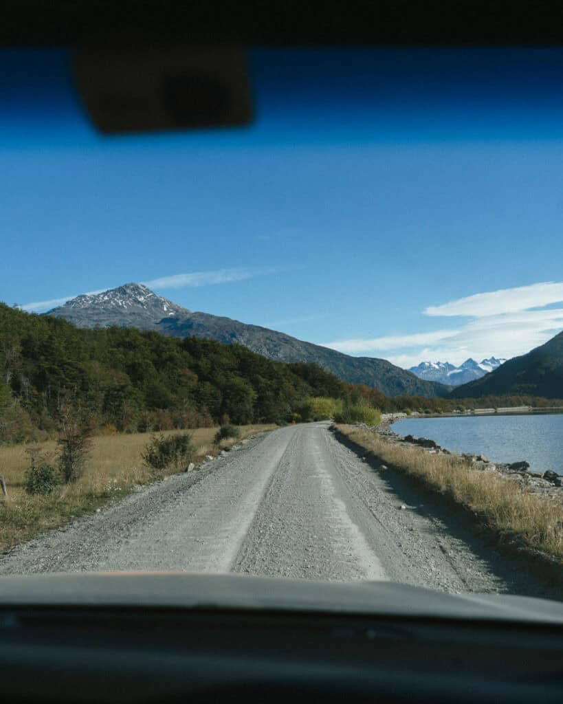 Villa O'Higgins, Chile Guide: End of the Carretera Austral 13 View from inside a vehicle heading down a remote gravel road next to a lake, with forested hills and distant snowy peaks near Villa O'Higgins