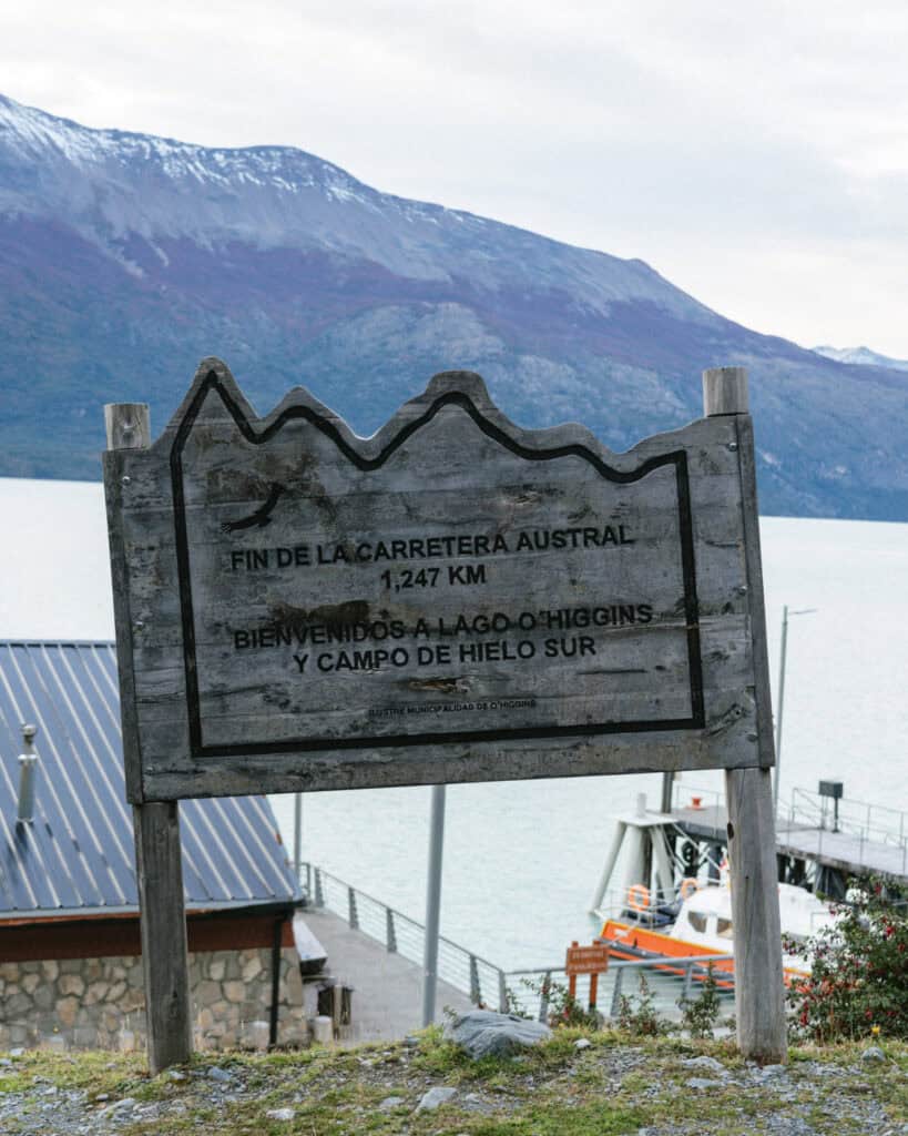 Villa O'Higgins, Chile Guide: End of the Carretera Austral 24 A rustic wooden sign reads "Fin de la Carretera Austral, 1,247 KM. Bienvenidos a Lago O'Higgins y Campo de Hielo Sur", positioned above a lakeside dock with boats and mountains in the background