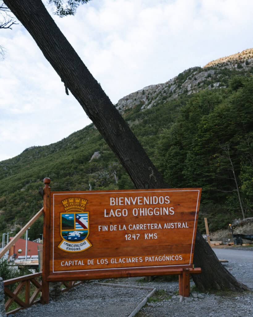 Villa O'Higgins, Chile Guide: End of the Carretera Austral 25 A polished wooden welcome sign in front of a forested mountain backdrop reads “Bienvenidos Lago O'Higgins - Fin de la Carretera Austral 1247 kms - Capital de los Glaciares Patagónicos"