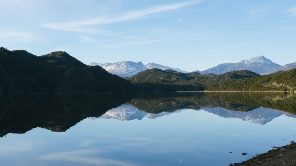 Villa O'Higgins, Chile Guide: End of the Carretera Austral 28 Crystal-clear reflection of snowy mountains and forested hills on the still waters of Lago Cisnes near Villa O'Higgins, with the moon faintly visible in the sky