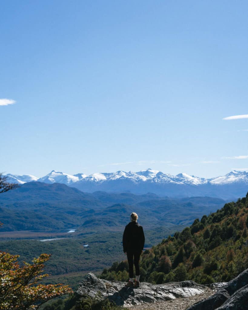 Camping on the Carretera Austral, Chile: Our Tips & Tricks 26 Brayden standing on a rocky outcrop looking out over an expansive green valley and distant snow-capped mountain range under a clear blue sky