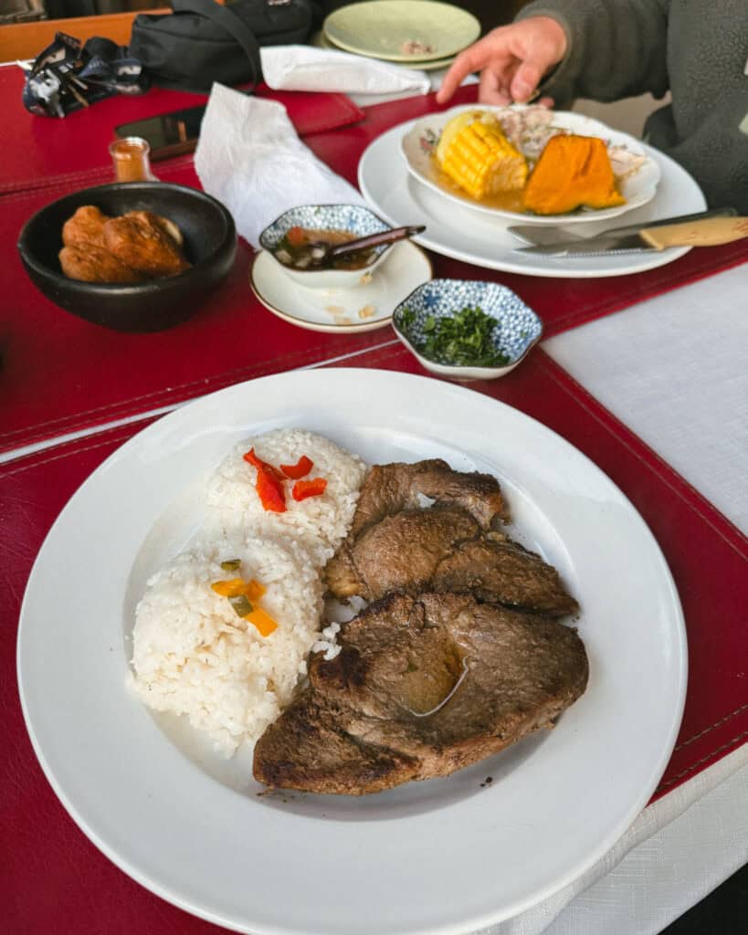Villa O'Higgins, Chile Guide: End of the Carretera Austral 30 A white plate with grilled beef slices served alongside two mounds of white rice, each topped with colorful pickled vegetables. Various side dishes including corn, pumpkin, and greens sit on a red table setting