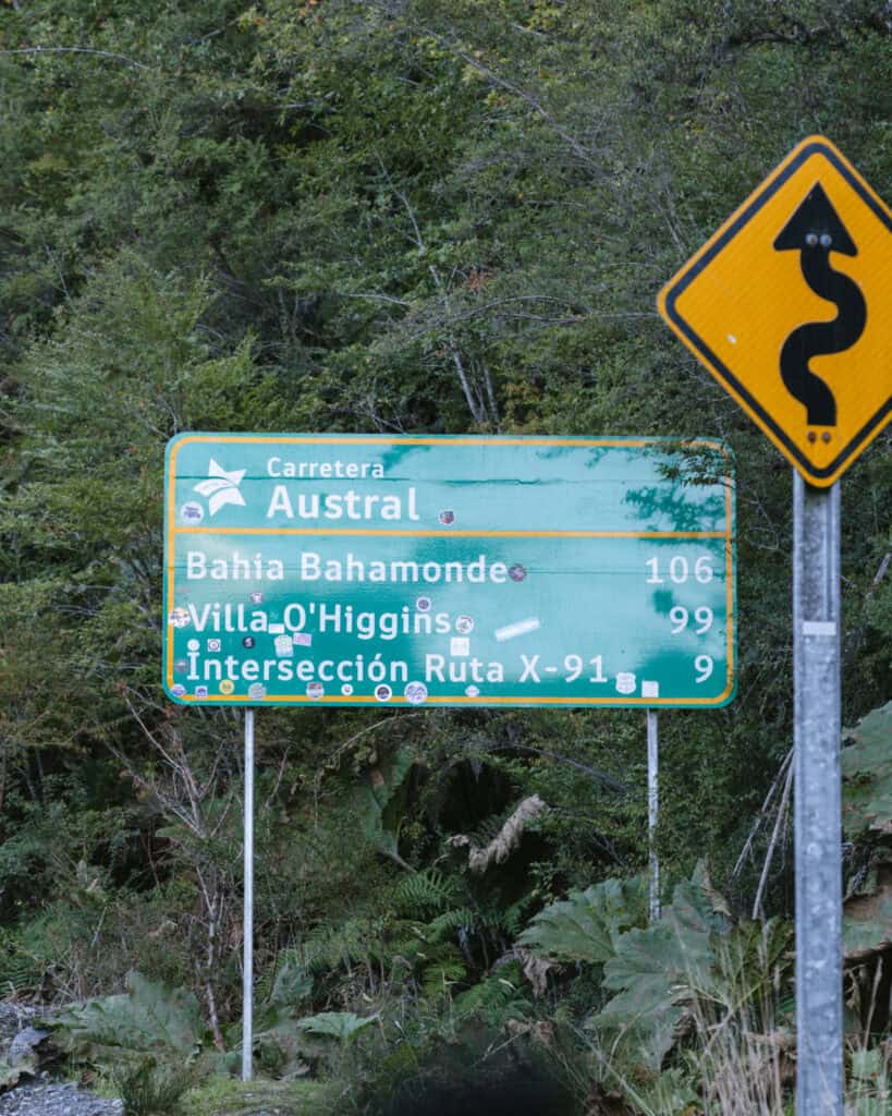 Villa O'Higgins, Chile Guide: End of the Carretera Austral 9 A green road sign along the Carretera Austral indicating distances to Bahia Bahamonde (106 km), Villa O'Higgins (99 km), and Ruta X-91 intersection (9 km), with a winding road caution sign beside it