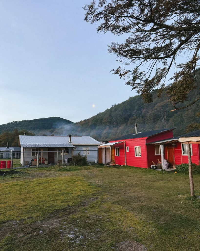 Villa O'Higgins, Chile Guide: End of the Carretera Austral 18 A rustic scene with a bright red and a pale blue house under a soft sky with the moon visible, and a trail of chimney smoke drifting up from one of the rooftops