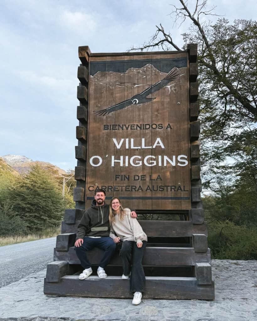 TABSA Ferry: Sailing from Puerto Natales to Puerto Yungay 47 Brayden and Sally sit on the base of a massive carved wooden sign that reads “Bienvenidos a Villa O'Higgins - Fin de la Carretera Austral,” framed by forest and mountains
