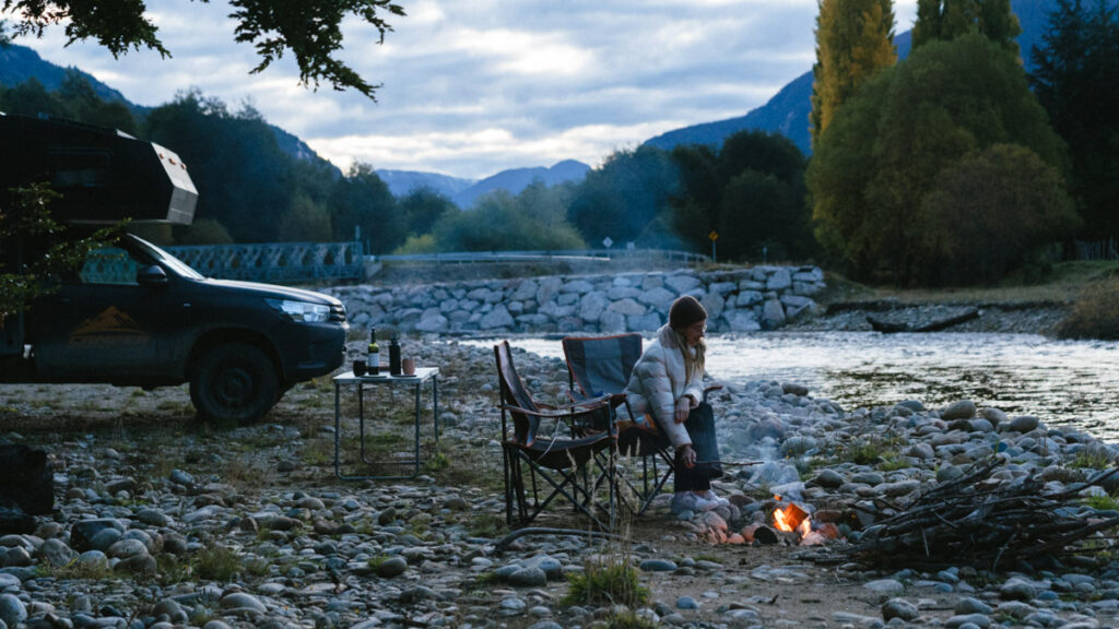 Camping on the Carretera Austral, Chile: Our Tips & Tricks 11 On a cloudy day in Villa Puente Piedra, Chile, Sally pokes a small fire beside a gently flowing river, with autumn trees and mountains in the distance