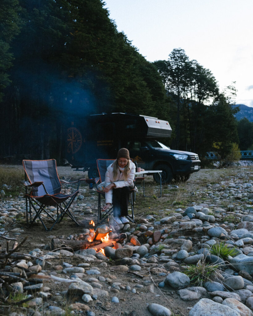 The Carretera Austral, Chile: Everything You Need to Know 66 Sally pokes at the campfire on a rocky shore in Villa Puente Piedra, Chile, framed by a forest backdrop and a camper van parked nearby