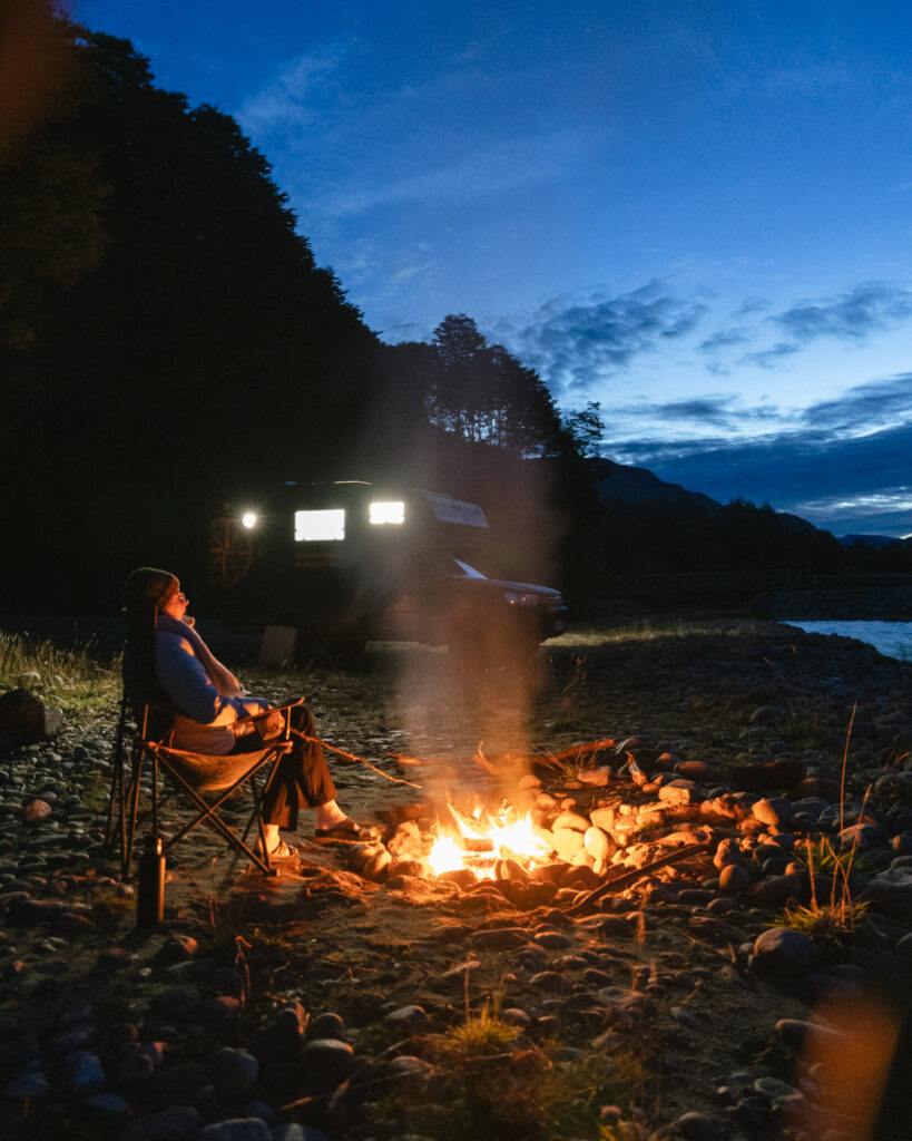 Camping on the Carretera Austral, Chile: Our Tips & Tricks 17 Seated by the fire in Villa Puente Piedra, Chile, Sally smiles while tending the flames, with pine trees and a black camper van in the background