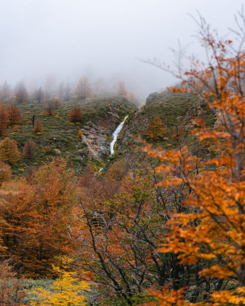 Laguna Cerro Castillo, Chile Hiking Guide 26 A thin waterfall cascades down a rocky hillside through a forest of vibrant orange and yellow trees, with soft mist hanging above the valley in Villa Cerro Castillo
