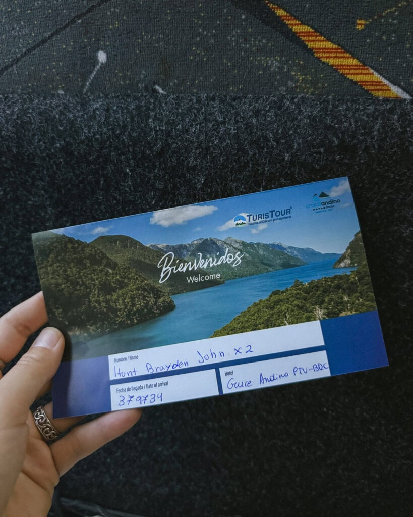 Cruce Andino: The Scenic Route From Chile to Argentina 7 A hand holding a welcome ticket from Turistour for the Cruce Andino experience in Chile, showing a scenic photo of a lake surrounded by lush green mountains. The handwritten name "Hunt Brayden John x2" and hotel "Guce Andino PTV-BDC" are visible