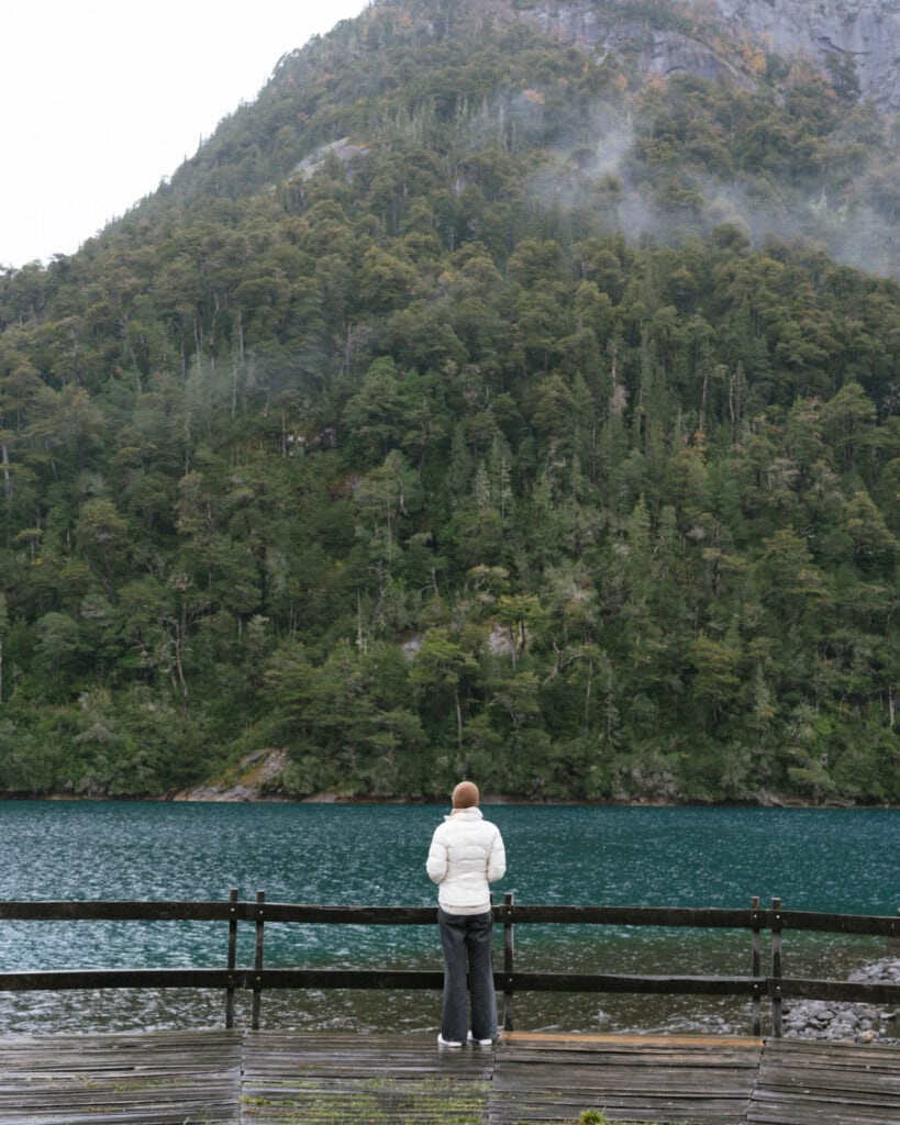 Cruce Andino: The Scenic Route From Chile to Argentina 29 Sally, wearing a white puffer jacket, stands on a wooden dock overlooking the calm turquoise waters of Puerto Blest, framed by a densely forested mountain