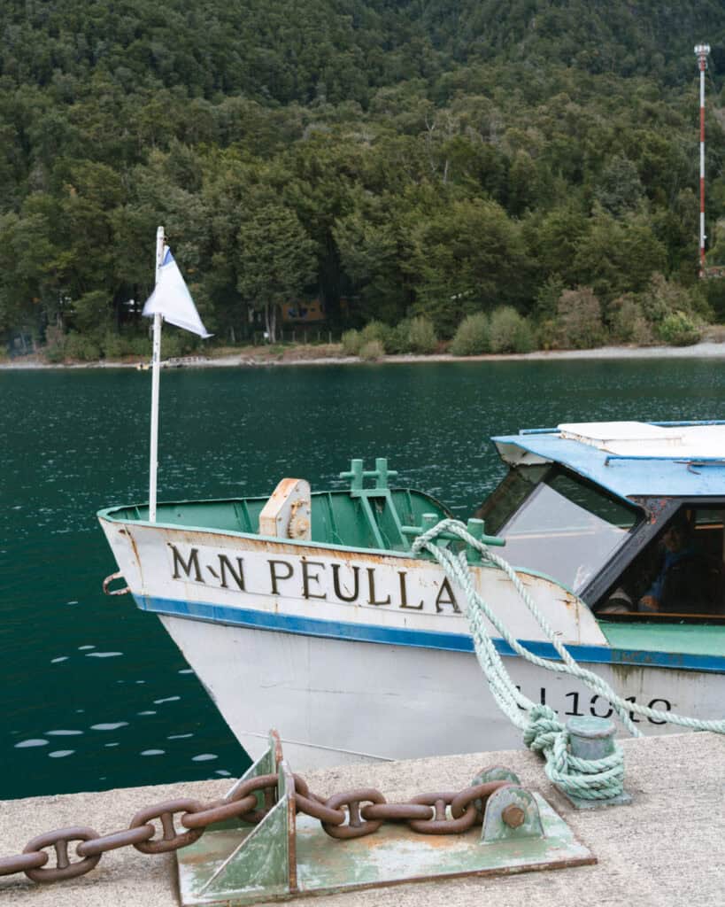 Cruce Andino: The Scenic Route From Chile to Argentina 13 The bow of a boat named “M.N. PEULLA” is tied to a dock on Lago Todos Los Santos, with emerald water and a thick tree line in the background