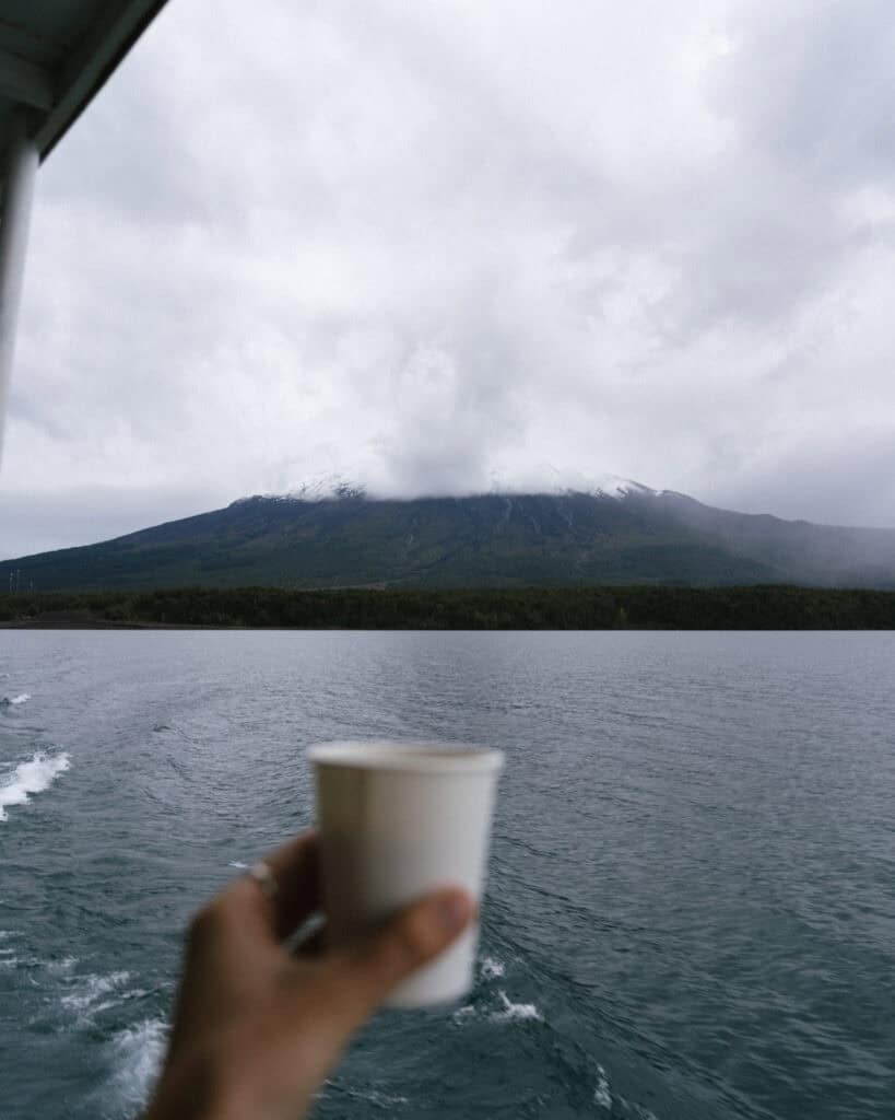 Cruce Andino: The Scenic Route From Chile to Argentina 17 A hand holds a white paper cup of coffee in the foreground, while a cloud-covered volcano and lush shoreline appear across Lago Todos Los Santos