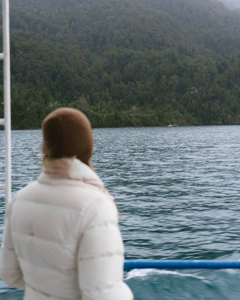 Cruce Andino: The Scenic Route From Chile to Argentina 16 Sally, wearing a white puffer jacket and brown beanie, stands on a boat deck looking out at the lush green hills and calm waters of Lago Todos Los Santos in Chile