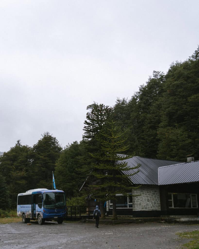 Cruce Andino: The Scenic Route From Chile to Argentina 23 A small blue Turistour bus is parked beside a rustic forest building near the Argentina-Chile border, with a hiker walking toward the entrance under a cloudy sky