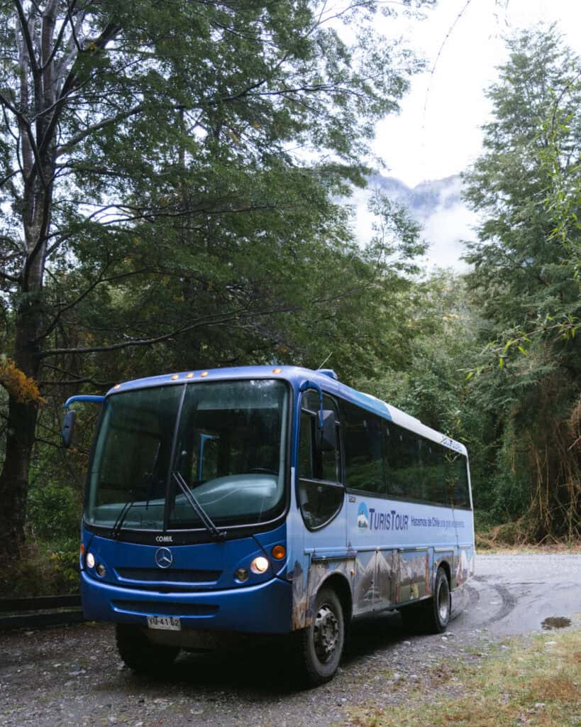 Cruce Andino: The Scenic Route From Chile to Argentina 19 A muddy blue Turistour bus stands on a gravel road surrounded by tall forest trees and mountain mist, near Peulla in the Chilean Andes