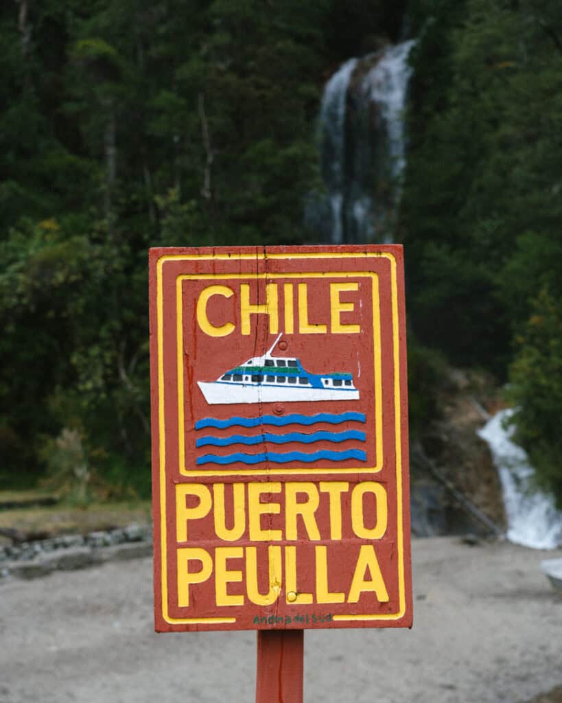 Cruce Andino: The Scenic Route From Chile to Argentina 18 A bright hand-painted wooden sign reading "Chile Puerto Peulla" stands in front of a cascading waterfall and lush forest, marking a key stop on the Cruce Andino route