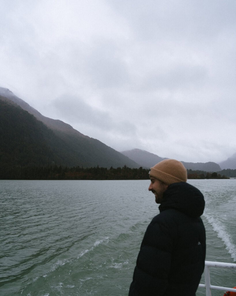 Cruce Andino: The Scenic Route From Chile to Argentina 27 Brayden smiles as he stands at the back of a boat cruising through Lago Frías, with green waters rippling behind him and misty Andean mountains rising in the background